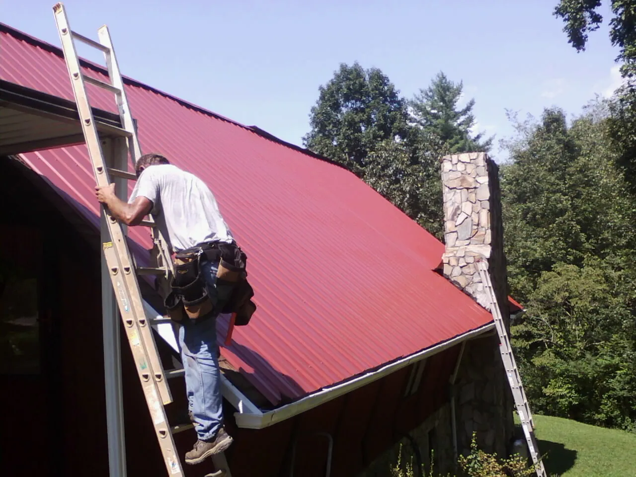 Roofer on a ladder beside a red metal roof and stone chimney — Soffit & Fascia Repair by Roofing Brothers in Glen Aubrey